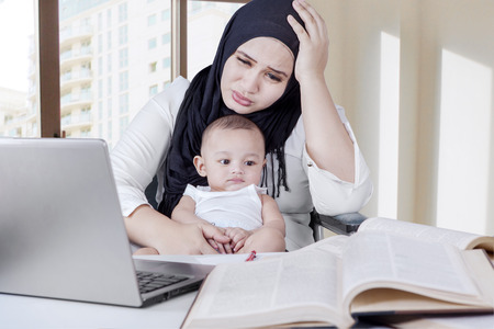 Young housewife looks tired, holding her baby while working on desk with laptop and booksの写真素材