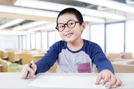 Male elementary school student sitting in the class while smiling at the camera and holding a marker with empty paperの写真素材
