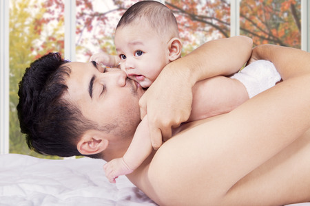 Young man lying on bed while holding and kiss his little baby, shot at home