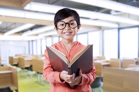 Portrait of beautiful little girl smiling on the camera while reading a book in the classroomの写真素材