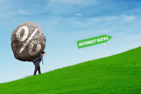 Businessman carries a stone with percentage rates symbol walking uphill following Interes Rates road signの写真素材
