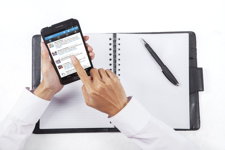 JAKARTA, SEPTEMBER 08, 2015: Closeup of businessman hand working with notebook while using a smartphone to open twitter site onlineのeditorial素材