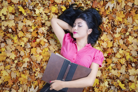 Portrait of female college student sleeping on the autumn leaves while holding a bookの写真素材