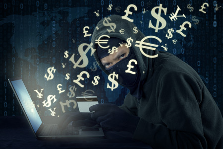 Image of male hacker wearing mask and using laptop while holding credit card to steal money through...