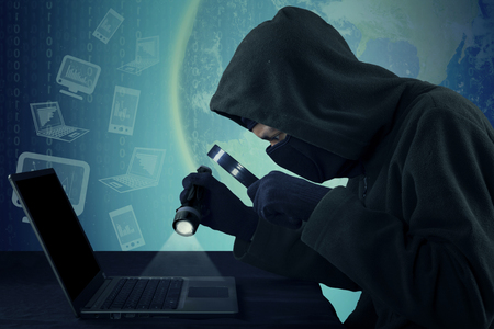 Photo of male thief wearing mask and steal user identity on the laptop while using flashlight and magnifying glassの写真素材