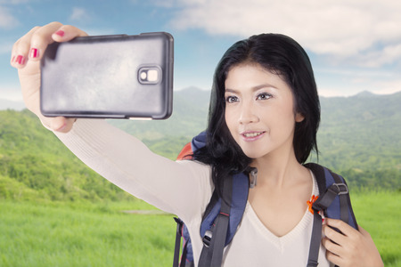 Portrait of pretty hiker carrying bag and holding smartphone to take selfie picture on the mountainの写真素材