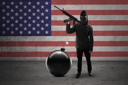 Concept of terrorism. Man wearing mask and holding weapon with bomb in front of American flagの写真素材