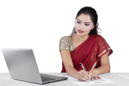 Portrait of indian businesswoman working with laptop while writing document on the paper and wearing traditional clothesの写真素材