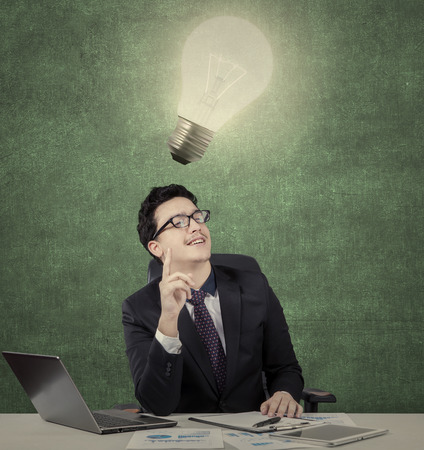 Middle eastern young businessman gets brilliant idea under bright lamp, shot with laptop computer and document on deskの写真素材
