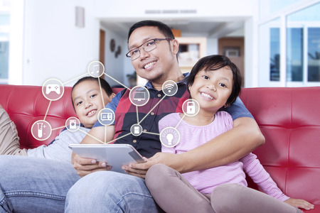 Happy father and his children sitting on the sofa while holding a digital tablet with smart house applicationsの写真素材