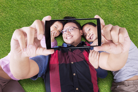 Two cheerful children lying on the park with their father and taking selfie photoの写真素材