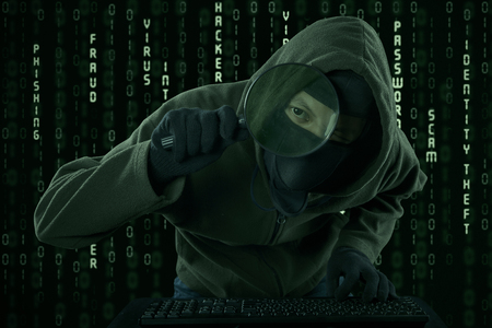 Internet Theft - a man wearing a balaclava looking at computer screen using magnifying glassの写真素材