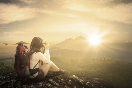 Back view of female traveler sitting on the rock while taking pictures with digital camera on the mountainの写真素材