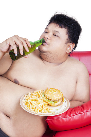 Portrait of young overweight man sitting on the sofa while enjoying fresh beer and burgerの写真素材
