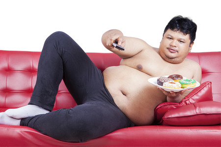 Photo of overweight man sitting on the sofa while holding a plate of donuts and remote, isolated on white backgroundの写真素材