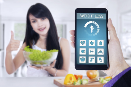 Close up of weight loss application on the mobile phone screen with young woman holding a bowl of...