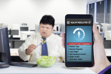 Image of hand holding smartphone with healthy resolutions program and overweight businessman eats salad in officeの写真素材