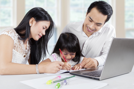 Two young Asian parents teach their daughter to write and doing homework with a laptop on desk at homeの写真素材