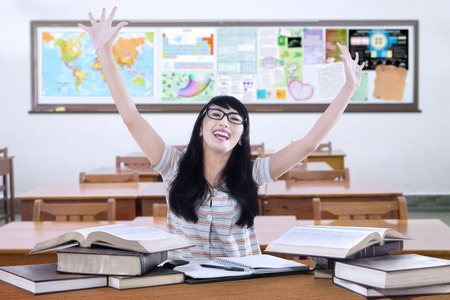 Portrait of happy female student raise hands in the classroom with books on the tableの写真素材