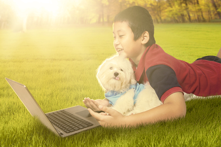 Image of happy little boy lying on the grass with his puppy while using laptop computer, shot on the parkの写真素材