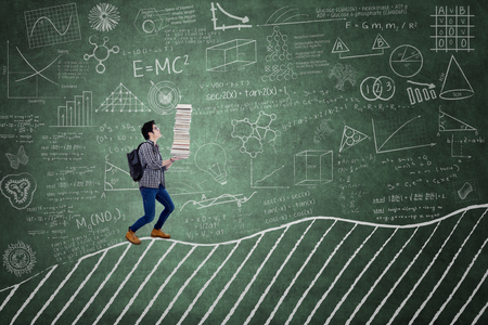 Picture of a male high school student carrying a stack of books on the chalkboard in the classroomの写真素材