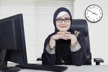 Picture of Arabic young woman wearing headscarf and smiling at the camera while sitting in the office with computer on the tableの写真素材