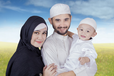 Two happy Arabic parents and their son smiling at the camera while wearing islamic clothes on the parkの写真素材