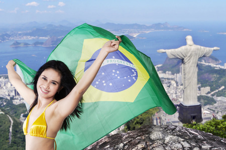 Portrait of cheerful young girl wearing bikini while holding flag of Brazil with Rio de Janeiro city backgroundのeditorial素材
