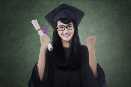 Beautiful female bachelor celebrating her graduation while holding certificate and wearing a mortarboard in classの写真素材