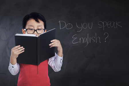 Little boy reading a book while wearing glasses and uniform with text Do You Speak English on the chalkboardの写真素材