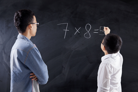 Little boy writes the answer of multiplication formula on the chalkboard with his teacher in the classの写真素材