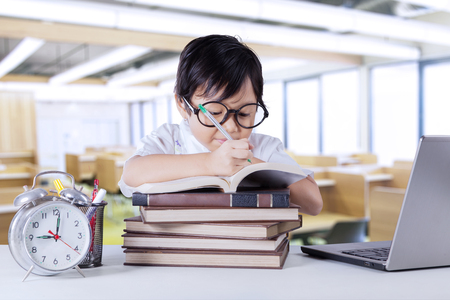 Female primary school student learns in the classroom while writing on the book with notebook computer and alarm clock on the tableの写真素材