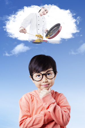 Pretty little girl wearing glasses and imagines a female chef cooking food on the thought bubbleの写真素材