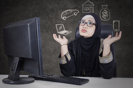 Arab business woman wearing glasses sitting in front of computer while doubting with her dream for buying something.の写真素材