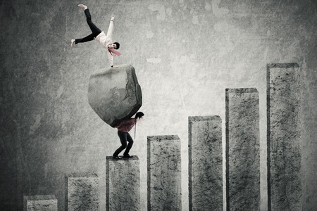 Image of happy businessman expressing success on a stone while his partner carrying the stone on the stairsの写真素材