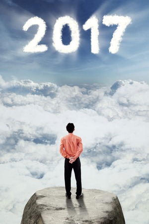Rear view of a businessman looking at number 2017 on the sky while standing on a rock, concept of a brighter future for his businessの写真素材