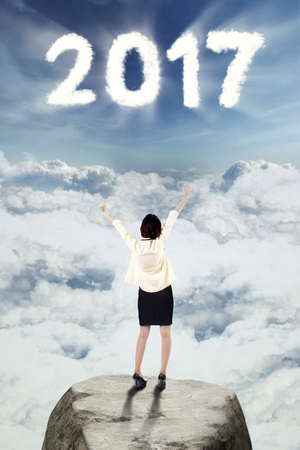 Back view of female entrepreneur standing on a rock while raising hands and looking at number 2017 on the skyの写真素材