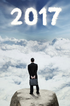 Image of male employee with a briefcase looking at number 2017 in the sky while standing on the rockの写真素材