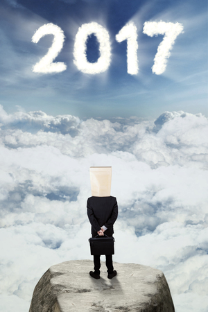 Male entrepreneur standing on a rock with number 2017 in the sky while carrying a briefcase and cardboard headの写真素材