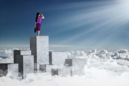 Image of a young businesswoman standing on the concrete wall and looking at blue sky using binocularsの写真素材