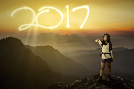 Image of beautiful backpacker woman showing thumb up and standing on the cliff with numbers 2017 on...