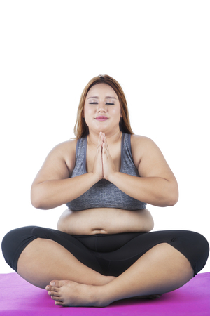 Image of overweight young woman sitting on a purple mattress while practicing yoga, isolated on white backgroundの写真素材