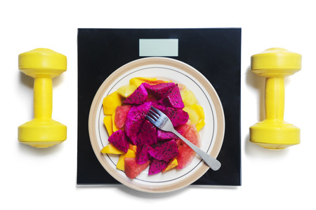 Top view of sliced fruits and fork on a plate with scales between yellow dumbbells, isolated on white backgroundの写真素材