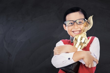 Portrait of a cheerful schoolboy smiling at the camera while wearing uniform and holding a trophy in the classの写真素材