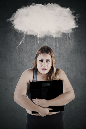 Depressed woman with overweight body holding a weighing scale under speech bubbleの写真素材