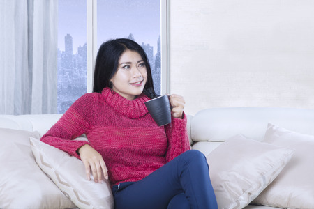Portrait of attractive woman holding a cup of hot coffee while sitting on the sofa with winter background on the windowの写真素材