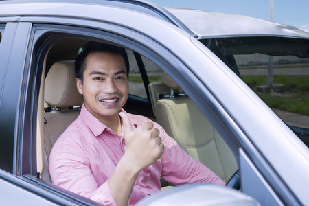 Image of happy young male driving a new car while showing thumb up and looking at the cameraの写真素材