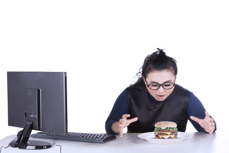 Portrait of female businesswoman looking at hamburger while sitting in front of computer, isolated on white backgroundの写真素材