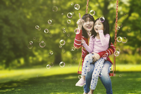 Portrait of young mother and cute child sitting on the swing while pointing at soap bubbles in the airの写真素材