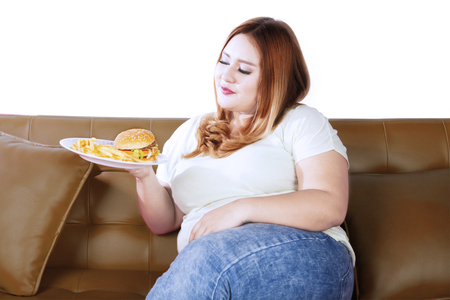 Image of obese woman holding junk food on a plate while sitting on the couch, isolated on white backgroundの写真素材